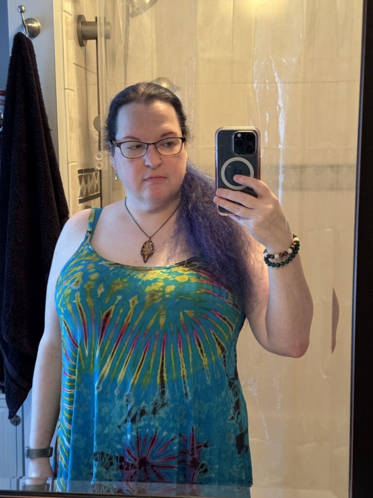 A mirror selfie of a white woman with purple hair in a ponytail, pulled over her shoulder. She's wearing a tie-dyed mostly blue tank top.