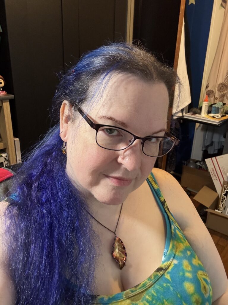 A selfie of the same woman sitting, with her head tilted to the side. The light brings out both the blue and purple in her hair, and she's wearing a copper leaf necklace.