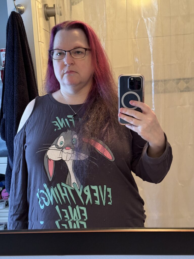 A mirror selfie of the same woman, hair loose and this time fading red to purple, wearing the same shirt. Her face is less round and the shirt is looser around her stomach.