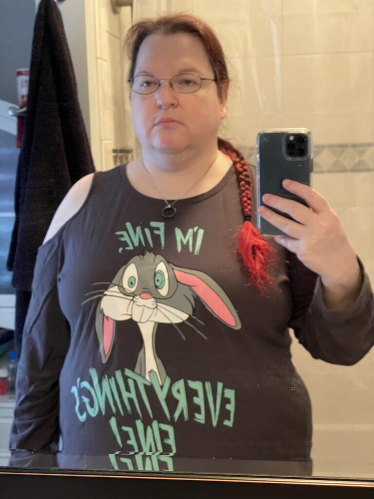 A mirror selfie of a fat white woman with glasses and bright red hair in a braid over her shoulder. She's wearing a long-sleeved cold shoulder gray shirt with a picture of Bugs Bunny with I'm fine, everything's fine written in blue lettering over and underneath him. The image contradicts the words.