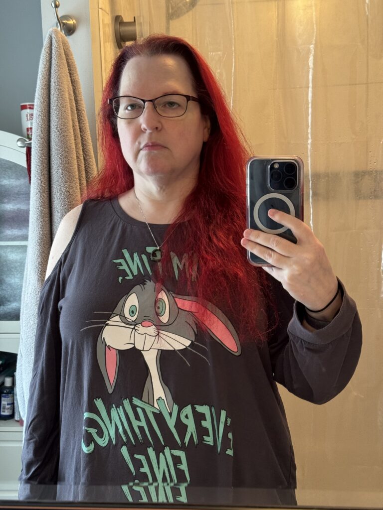 Another mirror selfie of the same woman, bright red hair loose and over her shoulder, wearing the same Bugs Bunny shirt. Her face is thinner, there's more definition on her neck, and the shirt hangs even looser.