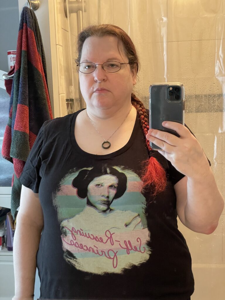 A mirror selfie of a fat white woman with red hair in a braid over her shoulder. She's wearing a Princess Leia shirt that says--backwards because mirrored--"Self-Rescuring Princess"